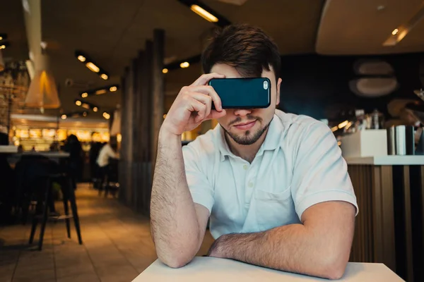 Retrato de un joven sentado en un restaurante y cubre sus ojos teléfono móvil — Foto de Stock