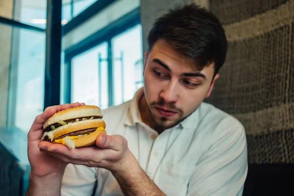 Młody człowiek siedzi w restauracji i przytrzymując mięso burger — Zdjęcie stockowe