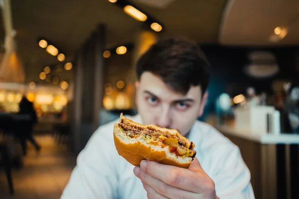 Estudiante con barba sentado en un bonito restaurante y ofrece una sabrosa hamburguesa — Foto de Stock