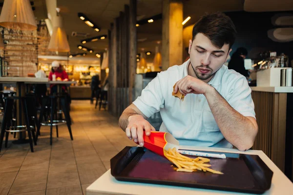 Молодой человек сидит в ресторане и быстро haruvannya наливает картошку фри в папку, чтобы затем поесть — стоковое фото