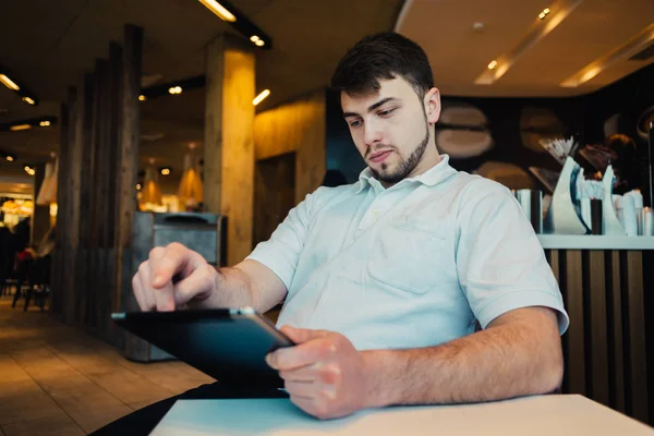 Ein junger Mann mit Bart sitzt in einem gemütlichen Café und benutzt einen Tablet-PC — Stockfoto