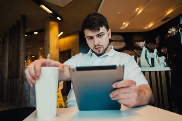 Un giovane studente di sesso maschile con un Tablet PC e una Coca-Cola fresca si siede in un tranquillo, bellissimo caffè — Foto Stock