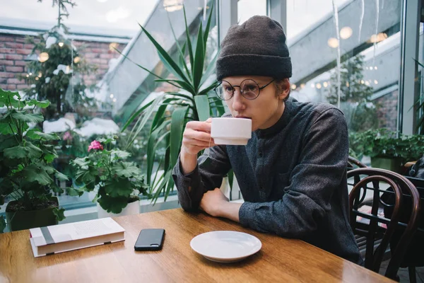 Un joven estudiante hipster bebe té en un café. tumbado en la mesa y la guía telefónica, y fuera del invernadero con plantas . — Foto de Stock