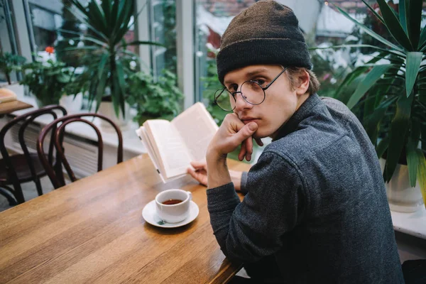 Joven estudiante sentado y bebiendo té y leyendo un libro en una cafetería durante un descanso en la universidad — Foto de Stock
