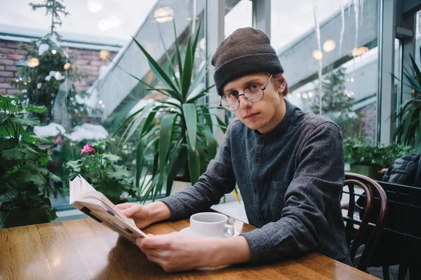Ο νεαρός άντρας hipster γυαλιά και καπέλο διαβάζοντας ένα ενδιαφέρον βιβλίο σε μια ωραία καφετέρια όπου πολύ πράσινο — Φωτογραφία Αρχείου