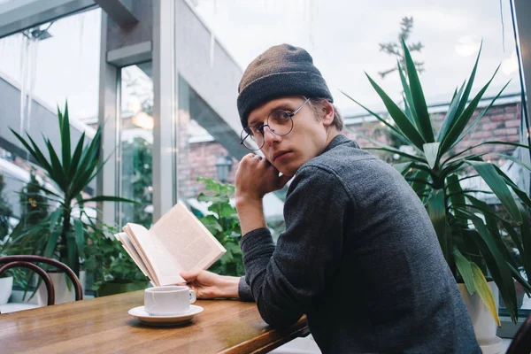 Молодой студент в очках, пьющий чай и читающий книгу в красивом кафе во время перерыва в университете — стоковое фото
