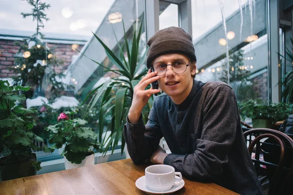 Giovane uomo in occhiali e una camicia che parla al telefono in un caffè con vista sul giardino d'inverno — Foto Stock