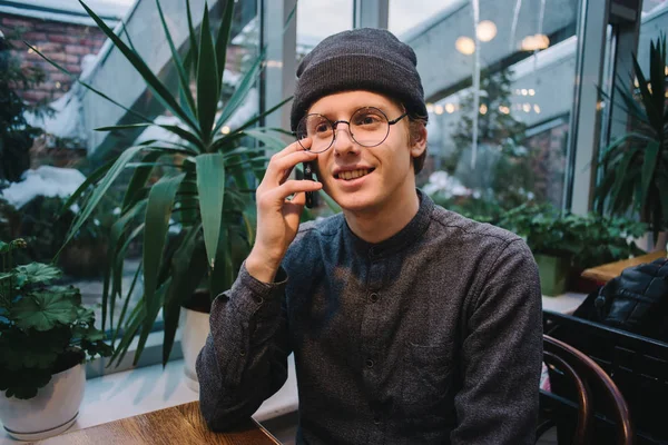 Счастливый молодой студент разговаривает по телефону у окна с видом на террасу с зелеными растениями — стоковое фото