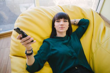 Genç güzel öğrenci eğitim ve bir telefon tutarak arasında dinleniyor. Bir odada bir koltuk çanta ile portre.