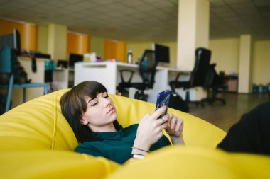 Bir sandalye çanta ve kullanım telefon oturma bir güzel genç ofis çalışanı portresi. Arka plan işleri karşı.