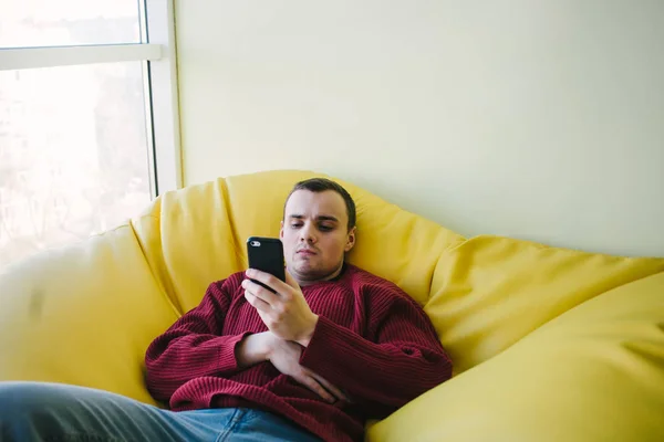 Der junge Mann sitzt als Büroangestellter in einem gelben Sesselsack und benutzt ein Smartphone. Pausenarbeit. — Stockfoto