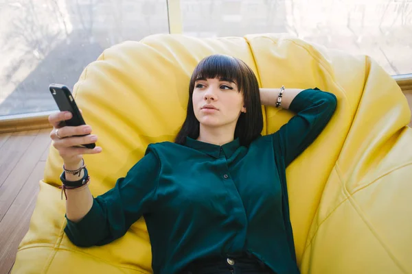 Genç güzel kız hipster geri yatıyor ve ışık cep telefonu sahiptir. Modern bir ofis sandalye çanta. Kapalı portre — Stok fotoğraf