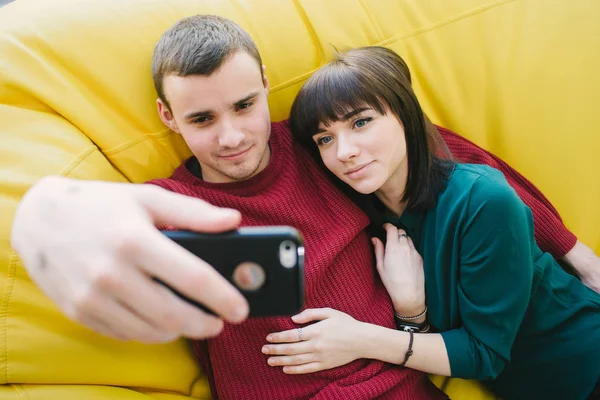 Dois jovens estudantes sorridentes fazem uma bela selfie. Retrato de jovens em um quarto em uma bolsa de cadeira amarela — Fotografia de Stock