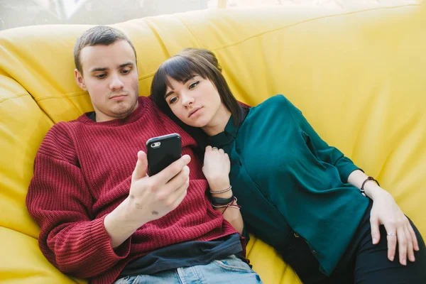 Jovem lindo casal apaixonado deitado em uma cadeira amarela e usar um telefone celular em seu tempo livre . — Fotografia de Stock