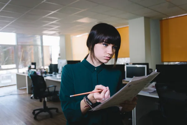 Joven chica hermosa oficinista mirando los papeles de negocios y escribe algo en un fondo de una oficina moderna . — Foto de Stock