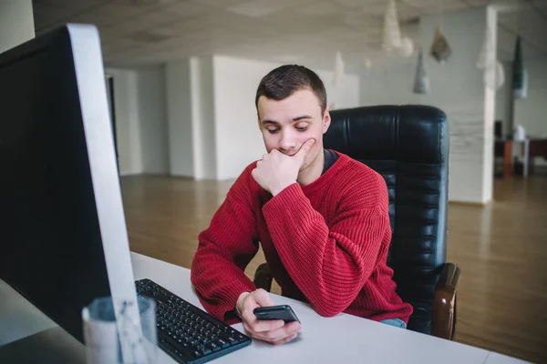 Joven diseñador hipster guapo sentado en el monitor de su computadora y utilizar un teléfono móvil. Vida de oficina . — Foto de Stock