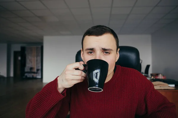 Retrato de un joven guapo sentado en el lugar de trabajo con una taza de café en la mano. Trabajador de oficina soñoliento — Foto de Stock