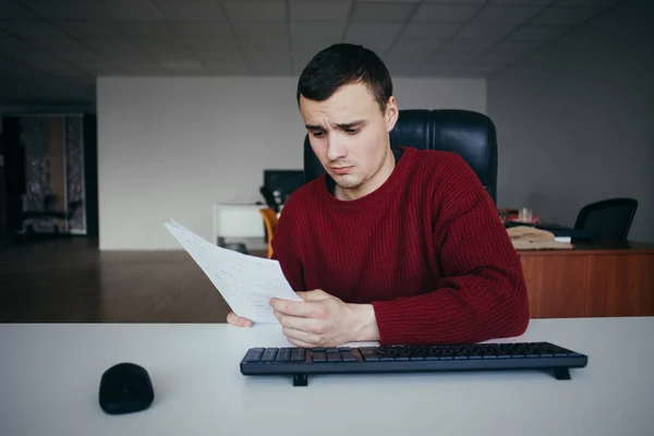 Молодий бізнесмен сидить на робочому місці і дивиться на папери в руках. На тлі офісу — стокове фото