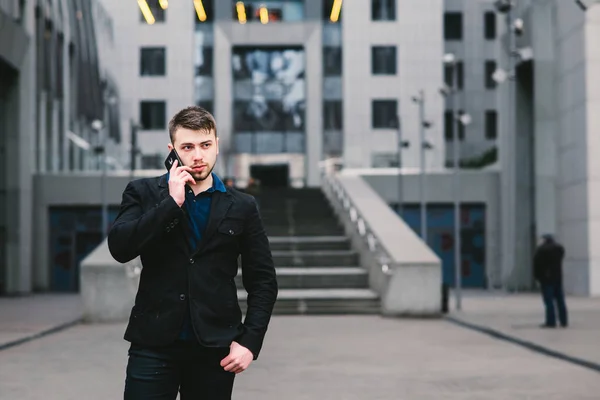 Joven hombre de negocios guapo en un traje oscuro y hablando por teléfono en el fondo de la arquitectura moderna — Foto de Stock