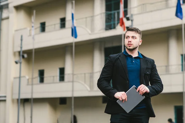 Joven empresario serio con una tableta en la mano en el fondo de la arquitectura y banderas de diferentes países . —  Fotos de Stock