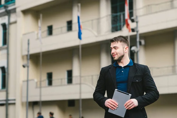 Joven hombre de negocios sonriente con tableta en la mano en el fondo de la arquitectura y banderas de diferentes países . —  Fotos de Stock