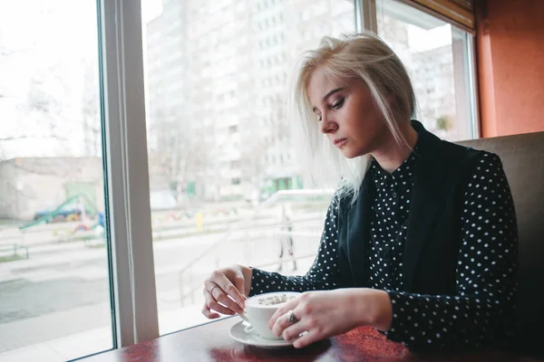 Jovem linda menina loira vestida de preto sentado no café com uma xícara de bebida quente.Café da manhã no café . — Fotografia de Stock