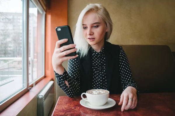 Menina loira jovem sentada em um café acolhedor com uma xícara de café e olhando atentamente para o telefone. Mulher marca uma mensagem em seu gadget — Fotografia de Stock