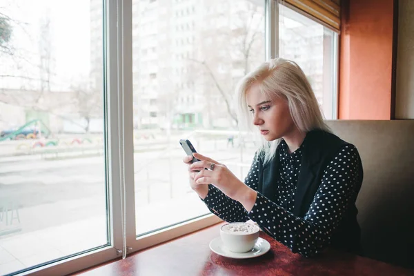 若い女子学生一杯のコーヒーと窓の近くの居心地の良いカフェに座っているとあなたの携帯電話でメッセージを得るします。コーヒー ブレーク — ストック写真