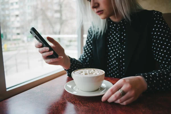 Μια νεαρή γυναίκα στο μαύρο χρησιμοποιεί το τηλέφωνο σε ένα καφέ και το ποτό καφέ με marshmallows. Διάλειμμα για καφέ στο καφενείο — Φωτογραφία Αρχείου