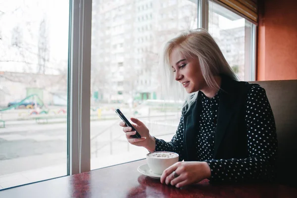 Молода дівчина посміхається блондинка дивиться в телефон, сидячи в кафе біля вікна з чашкою гарячого напою. Ранкова чашка кави зі студентом — стокове фото
