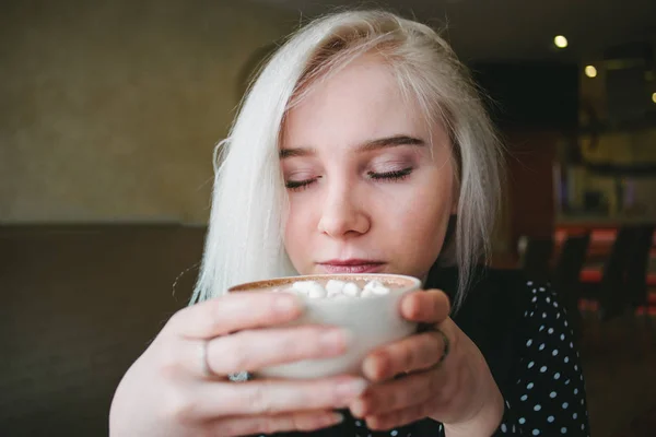Retrato de una joven rubia hermosa que disfruta de una taza de café con malvaviscos en un café. Mujer con los ojos cerrados disfrutando del aroma del café — Foto de Stock