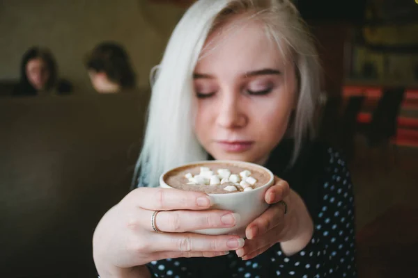 Joven hermosa chica rubia disfruta de una taza de cacao con malvaviscos y un acogedor café . — Foto de Stock