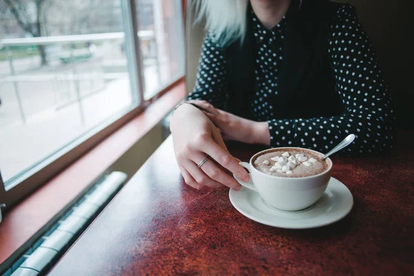 Uma mulher a segurar uma bebida quente. Uma xícara de café com marshmallows em um café na mesa e mãos a mulher vai beber. Cacau com marshmallows . — Fotografia de Stock