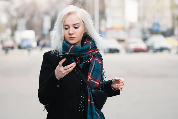 Όμορφη νεαρή ξανθιά ντυμένοι με ζεστά ρούχα, το περπάτημα κάτω από την οδό και κοιτάζοντας το κινητό τηλέφωνο. πορτραίτου. — Φωτογραφία Αρχείου