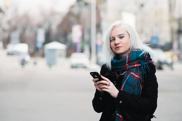 Πορτραίτου του μια όμορφη κοπέλα στον µαθητή ρούχα φθινόπωρο που χρησιμοποιεί ένα τηλέφωνο για το φόντο της πόλης. Ξανθιά μέση του δρόμου με το τηλέφωνο στο χέρι — Φωτογραφία Αρχείου