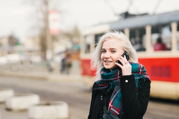Όμορφη ξανθιά κοπέλα φορώντας ζεστό ότι μιλάμε στο τηλέφωνο για το φόντο του αστικού τοπίου. Πορτραίτου για το φόντο του τραμ — Φωτογραφία Αρχείου