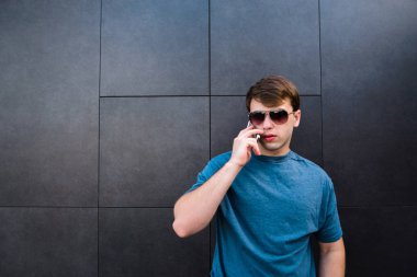 Yakışıklı bir genç adam gözlük ve bir T-shirt telefonla gri bir duvar arka plan üzerinde konuşuyor..