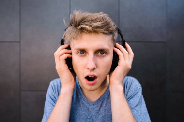 Komik bir genç adam şaşırmış gibi yüzü olan bir arka plan gri duvar Kulaklığımda müzik dinler. Duygusal genç müzik zevk alır. Kameraya bak.