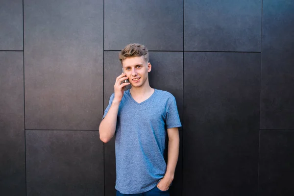 Um jovem sorridente em uma camiseta cinza fala com amigos em um telefone celular contra um fundo de parede cinza. Conversas de telefone celular com amigos . — Fotografia de Stock