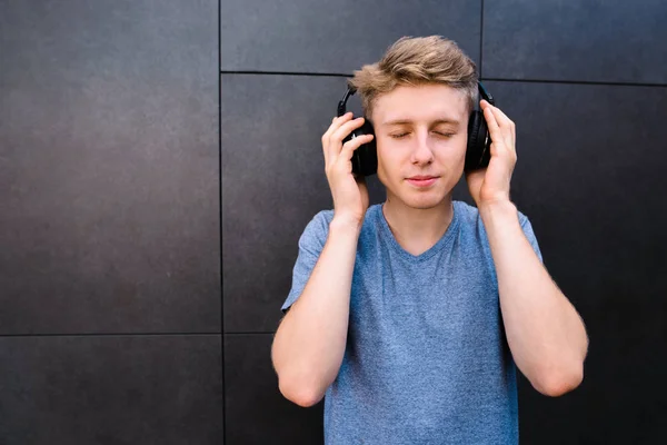 Красивый молодой человек с закрытыми глазами слушает музыку в наушниках на фоне серой стены. Подросток наслаждается музыкой. — стоковое фото