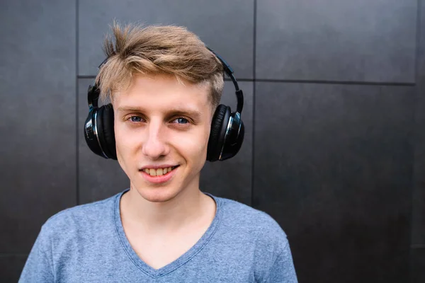 Portrait of a smiling beautiful man in a wireless headphones on the background of the wall. A positive teen gets fun with listening to music. — Stock Photo, Image