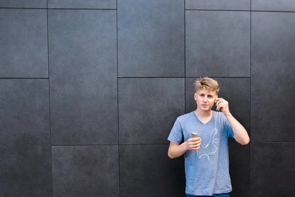 Estudante em uma camiseta cinza fica com um telefone e mãos no fundo da parede e insere um fone de ouvido no ouvido . — Fotografia de Stock