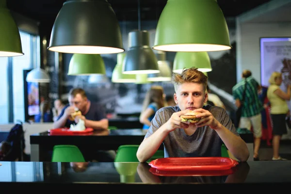 El joven se sienta en un restaurante de comida rápida con una hamburguesa en sus manos. Deliciosa comida para el almuerzo . — Foto de Stock