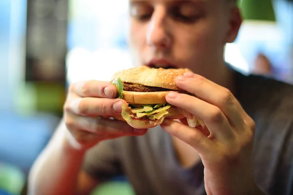 학생은 패스트 푸드 레스토랑에 앉아 위 대 하 고 맛 있는 햄버거를 먹을 것 이다. 햄버거에 초점은, 젊은 사람이 흐리게. — 스톡 사진