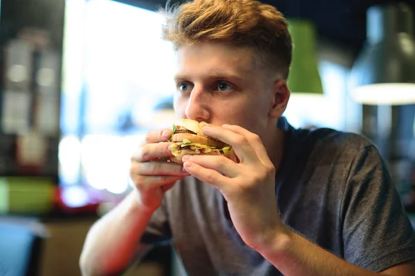 Un estudiante come una deliciosa hamburguesa sentada en un restaurante de comida rápida. Pausa para el almuerzo con mala comida . — Foto de Stock