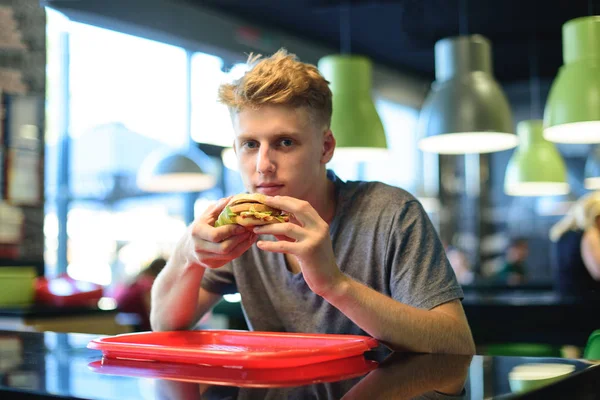 A student eats fast food in a bistro. An appetizing burger in the hands of a young man. A look at the camera.