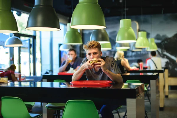 Joven estudiante hermosa sentado en un restaurante de comida rápida y comer una hamburguesa apetitosa . — Foto de Stock