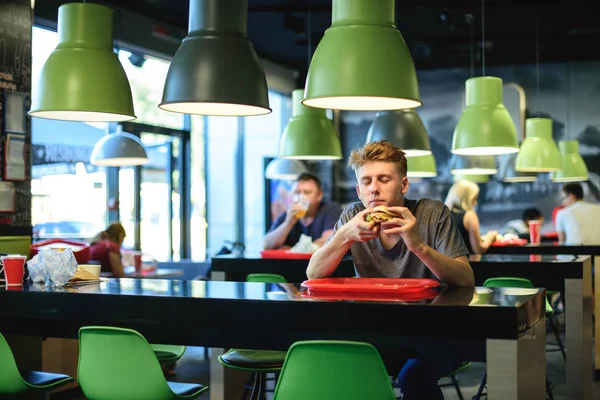 El joven y guapo hombre con los ojos cerrados disfruta de una hamburguesa en un restaurante de comida rápida . — Foto de Stock