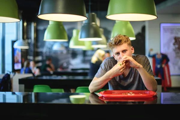 Um jovem gananciosamente come um hambúrguer apetitoso sentado em um restaurante de fast food . — Fotografia de Stock