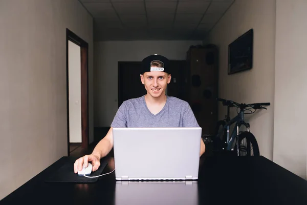Улыбающийся красивый студент в кепке и футболке пользуется ноутбуком дома за столом — стоковое фото